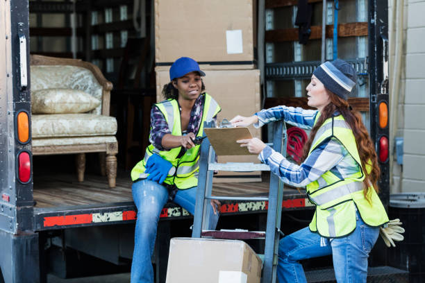 istockphoto-930527976-612x612 Two multi-ethnic female workers in their 30s and 40s at the back of a truck, one holding a clipboard. The women are wearing reflective vests and jeans. They are moving merchandise for a furniture store.